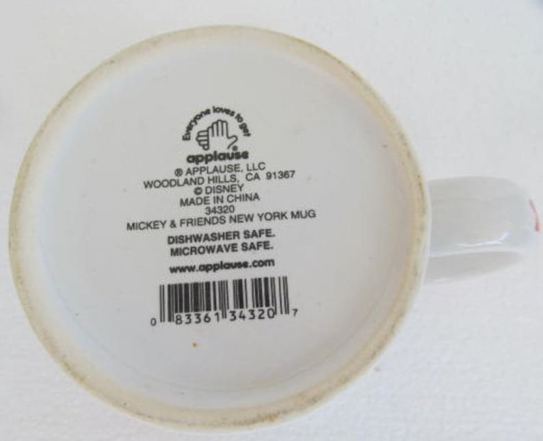 May include: White ceramic mug with a black and white image of Mickey Mouse and the text "Everyone loves to get applause" on the front. The back of the mug has the text "Applause, LLC Woodland Hills, CA 91367 &copy; Disney Made in China 34320 Mickey & Friends New York Mug Dishwasher Safe. Microwave Safe. www.applause.com" and a barcode.