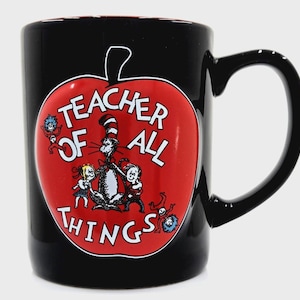 May include: Black ceramic mug featuring a red apple design with the text "Teacher of All Things." The apple graphic includes illustrations of characters from a children's book. The mug has a black handle and interior.