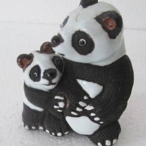 Puede incluir: Una figurita de cerámica de una madre panda sosteniendo a su bebé panda. La madre panda es negra y blanca con orejas marrones y una nariz negra. El bebé panda también es negro y blanco con orejas marrones y una nariz negra.