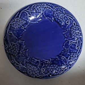 May include: A blue ceramic plate with a raised design of grape vines and grapes around the rim.