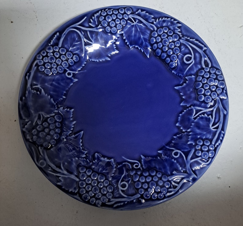 May include: A blue ceramic plate with a raised design of grape vines and grapes around the rim.