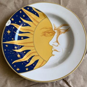 May include: A decorative plate with a celestial design. The plate has a white base and a gold rim. One half features a yellow sun with a face, the other a dark blue sky with stars and a crescent moon. The plate is likely ceramic.
