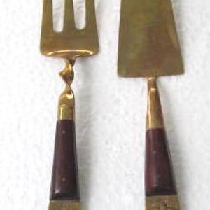 May include: A set of two brass serving utensils with wooden handles. The fork has a twisted handle and the knife has a rounded blade. Both utensils have decorative designs on the handles.