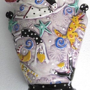 May include: A purple ceramic wall vase with a red, white and black rim. The vase is decorated with a whimsical design of a woman flying, a dog and stars. The vase has a black base with white polka dots.