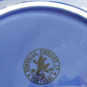 May include: Close-up of the bottom of a blue ceramic plate with a black logo that reads "Bordallo Pinheiro Lda Portugal" and features a frog silhouette.