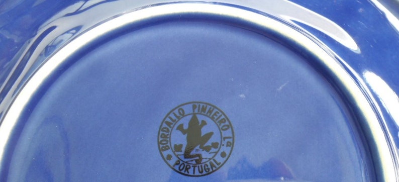 May include: Close-up of the bottom of a blue ceramic plate with a black logo that reads "Bordallo Pinheiro Lda Portugal" and features a frog silhouette.