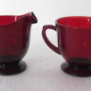 Può includere: Un set di due brocche per la panna e zuccheriera in vetro rosso vintage. La brocca per la panna ha un solo manico e un beccuccio. La zuccheriera ha due manici.