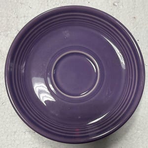 May include: A purple ceramic saucer with concentric rings and a central recessed area. The saucer has a glossy finish and a simple, functional design. This dish is suitable for a teacup or small cup.