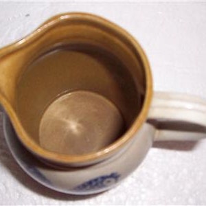 May include: A small, cream-colored ceramic pitcher with a brown glaze on the inside and a blue and white floral design on the bottom. The pitcher has a handle and a spout.