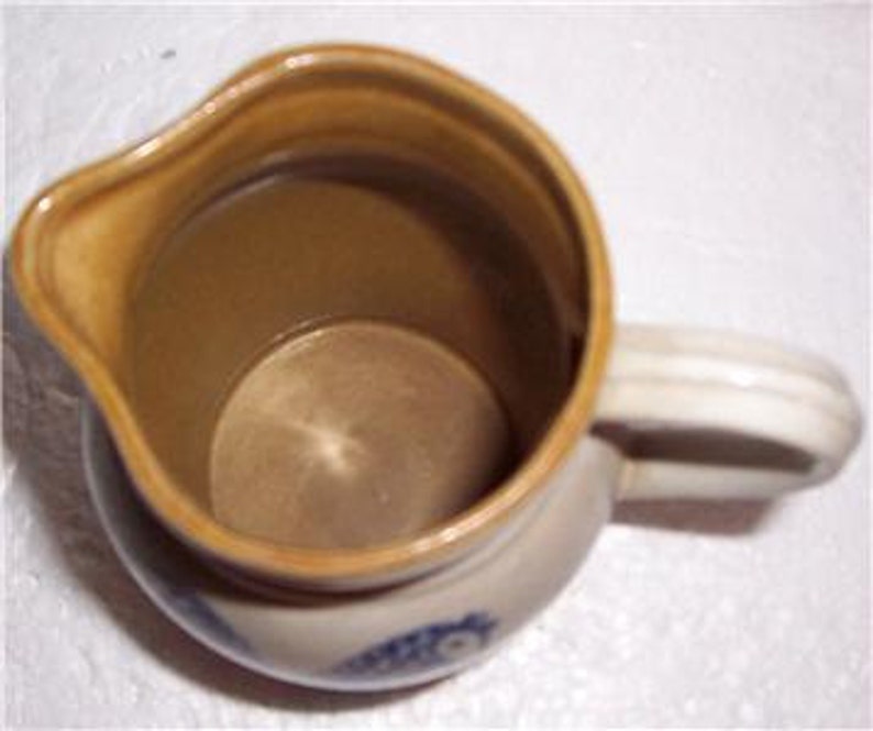 May include: A small, cream-colored ceramic pitcher with a brown glaze on the inside and a blue and white floral design on the bottom. The pitcher has a handle and a spout.