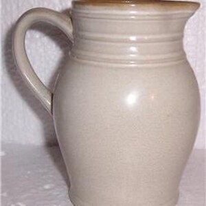 May include: A cream-colored ceramic pitcher with a brown rim and three horizontal lines around the body. The pitcher has a rounded body and a handle.