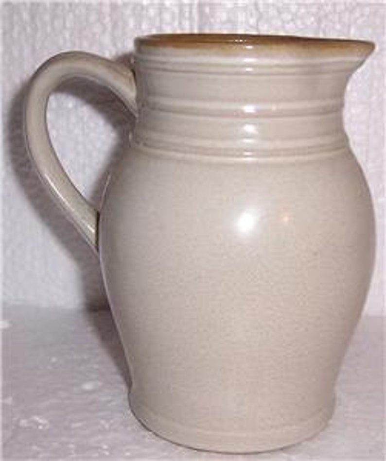May include: A cream-colored ceramic pitcher with a brown rim and three horizontal lines around the body. The pitcher has a rounded body and a handle.