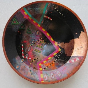May include: A round ceramic bowl with a black background and colourful abstract designs. The bowl features geometric shapes, stripes, and swirls in shades of pink, yellow, red, blue, and green. The bowl has a copper-coloured rim.