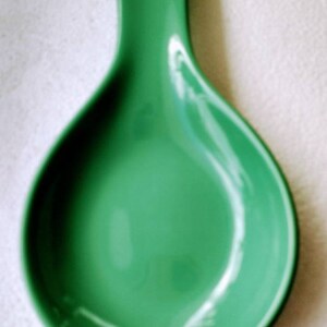 May include: A green ceramic spoon rest with a smooth, glossy finish. The rest has a curved shape with a wide, shallow bowl.