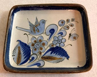 Könnte beinhalten: Quadratischer Keramikteller mit blauem und braunem Blumen- und Vogeldesign. Der Teller hat einen cremefarbenen Hintergrund mit einem blauen Rand. Das Design umfasst einen blauen Vogel, Blumen und einen Schmetterling.