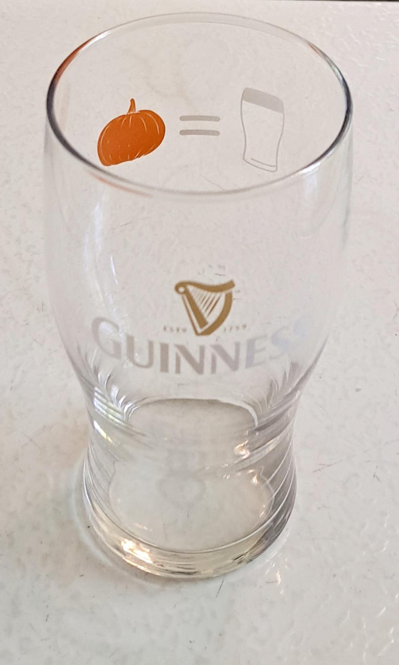 May include: A clear Guinness pint glass with a pumpkin graphic and the equation "pumpkin = beer". The Guinness logo and harp are visible. The glass is designed for beer.