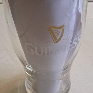 May include: Clear pint glass with the Guinness logo, featuring a gold harp and the word "Guinness" in white. The glass is partially filled with white paper. The text "ESTD 1759" is also visible.