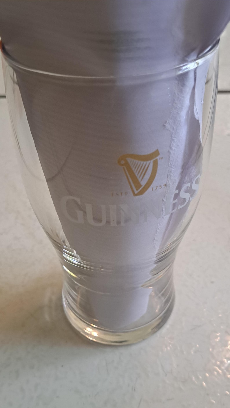 May include: Clear pint glass with the Guinness logo, featuring a gold harp and the word "Guinness" in white. The glass is partially filled with white paper. The text "ESTD 1759" is also visible.