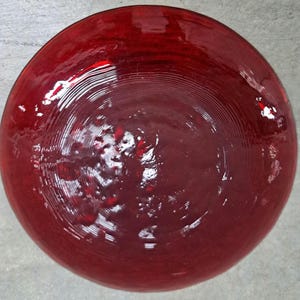 May include: A round, deep red glass bowl with a glossy finish. The bowl has a series of concentric circular ridges radiating from the center. The bowl's color is a rich, saturated red, and it reflects light.