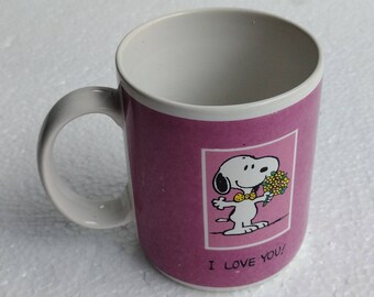Può includere: Una tazza in ceramica rosa con un'immagine di Snoopy dei cartoni animati che tiene un mazzo di fiori gialli. La tazza ha un manico bianco e il testo "I love you!" stampato sul fondo.