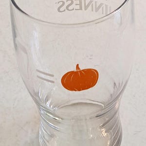 May include: Clear glass Guinness pint glass with a pumpkin graphic. The glass has the Guinness logo and the word "GUINNESS" printed in silver. The pumpkin is orange and is located on the front of the glass.