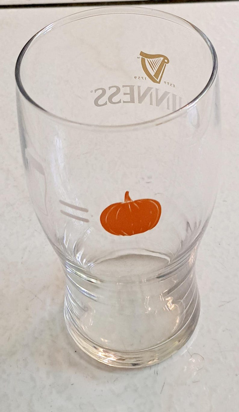 May include: Clear glass Guinness pint glass with a pumpkin graphic. The glass has the Guinness logo and the word "GUINNESS" printed in silver. The pumpkin is orange and is located on the front of the glass.