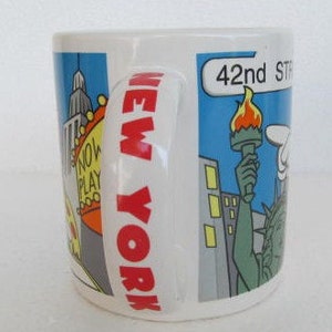 May include: A white ceramic mug with a red "NEW YORK" graphic on the handle. The mug features a cartoon illustration of New York City landmarks, including the Empire State Building, a yellow taxi, a pizza slice, and the Statue of Liberty. The text "42nd STREET" is visible on the mug.
