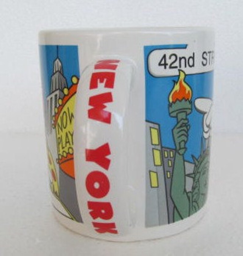 May include: A white ceramic mug with a red "NEW YORK" graphic on the handle. The mug features a cartoon illustration of New York City landmarks, including the Empire State Building, a yellow taxi, a pizza slice, and the Statue of Liberty. The text "42nd STREET" is visible on the mug.