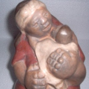 Puede incluir: Una escultura de cerámica de una mujer sosteniendo a un niño. La mujer lleva un vestido rojo y tiene una cubierta de cabeza blanca. El niño está envuelto en una tela blanca y tiene la cara marrón.