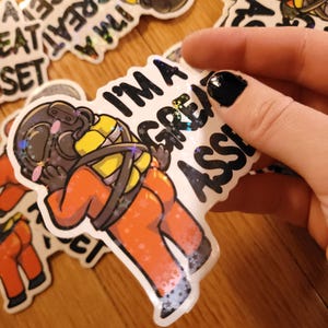 May include: Holographic sticker featuring a cartoon diver in an orange suit, with the text "I'M A GREAT ASS-ET". The sticker has a shiny, iridescent finish with star-shaped accents. Other stickers are visible in the background.