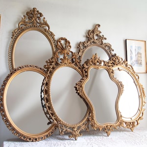 May include: A collection of five gold-toned mirrors with ornate, decorative frames. The mirrors vary in shape, including oval, circular, and irregular forms, all featuring intricate scrollwork and detailed carvings along their edges.