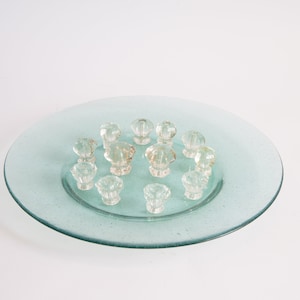 May include: A clear glass plate with a textured surface, holding a collection of clear glass knobs. The knobs are arranged in a circular pattern on the plate.