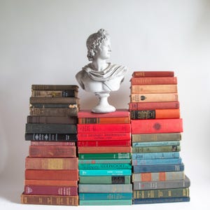 May include: A white plaster bust of a classical figure sits atop a stack of vintage books. The books are in various colors, including red, green, and brown. The books are arranged in a tiered display.