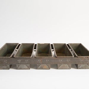 May include: A vintage metal loaf pan with six individual compartments. The pan is made of a dark gray metal and has a slightly worn appearance. The pan is labeled "Kellogg's" on the bottom.