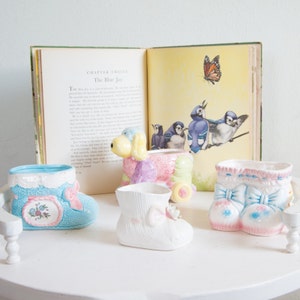 May include: Four ceramic planters shaped like a blue shoe, a white shoe, a pink and blue shoe, and a pastel-colored poodle. The planters are sitting on a white surface with a book open to a page about the blue jay.