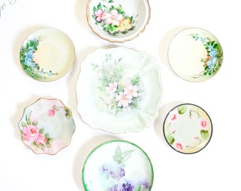 May include: A collection of seven vintage porcelain plates with floral designs. The plates are in various sizes and shapes, and feature different floral patterns, including roses, forget-me-nots, and other delicate blooms. The plates are all white with gold trim.