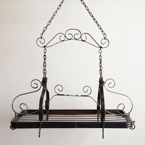 May include: Black wrought iron hanging pot rack with a rectangular shelf and decorative scrollwork. The rack is suspended by chains and features a wooden base. A stylish kitchen storage solution.