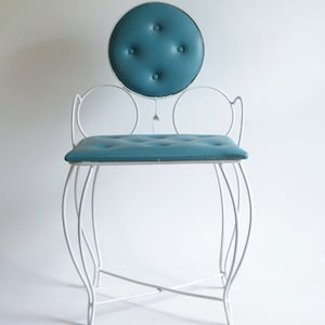 May include: A vanity chair featuring a white metal frame with curved legs and circular armrests. The seat and the circular backrest are upholstered in a teal blue material with button tufting. The backrest is supported by a single metal post.