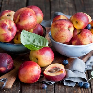 May include: Two bowls filled with ripe peaches, one bowl is blue and the other is white. The peaches are arranged in a cluster and have a red and yellow skin. There are blueberries scattered around the bowls on a wooden surface.