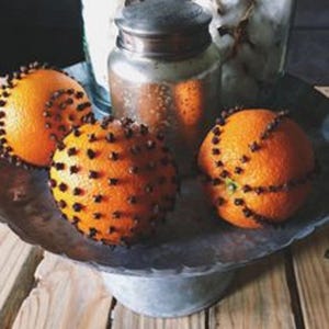 May include: Three oranges studded with cloves, arranged on a rustic metal tray. The oranges are a vibrant orange color and the cloves are dark brown. The tray is a dark gray color and has a rough texture.