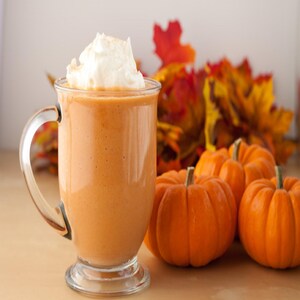 May include: A glass mug filled with a pumpkin spice smoothie topped with whipped cream. The mug is sitting on a table with small orange pumpkins and fall leaves in the background.