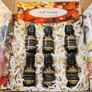 May include: A gift box filled with six small brown glass bottles of essential oils. The bottles are labeled with gold lettering and feature fall-themed scents like Apple Harvest, Pumpkin Spice, and Apple Maple Bread. The box is decorated with a fall-themed design and the text "Fall Harvest" and "Premium Home Fragrance Oils".