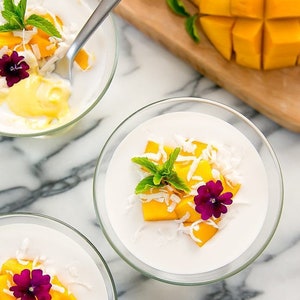May include: Three clear glass bowls filled with a white creamy dessert topped with diced mango, shredded coconut, and a sprig of mint. Each bowl is decorated with a single purple flower.