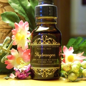 May include: A small glass bottle with a black label that reads "Riley Creek Floating Candles Pandora, Ohio Hydrangea 100% Homemade Soy Candle 419-384-7150 RileyCreekFloatingCandles.com". The bottle is surrounded by pink and white flowers.