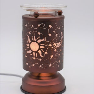 May include: A bronze-colored electric essential oil diffuser with a celestial design. The cylindrical body features cut-out stars, a sun, and a crescent moon. A clear glass dish sits atop the diffuser, and a power cord extends from the base.