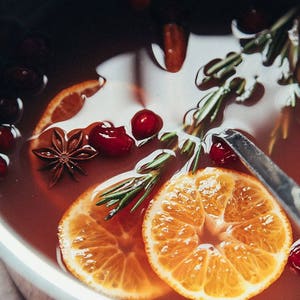 May include: A close-up of a pot filled with a red liquid, orange slices, cranberries, rosemary sprigs, and star anise.