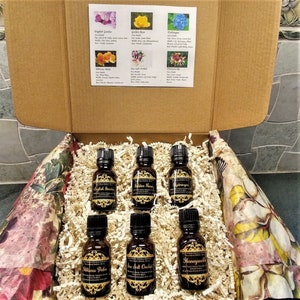May include: A box filled with brown packing paper and six small glass bottles with gold labels. The labels have names of different floral scents, including English Garden, Golden Rose, Hydrangea, Hibiscus Palm, Sea Salt Orchid, and Honeysuckle.
