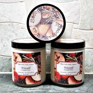 May include: Two glass jar candles with black lids and labels featuring apples and rosemary. The labels read "Wassail" and "Scoopable Wax". A circular label with the words "Scoopable Wax Wassail" is on top.