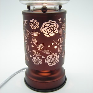 May include: A decorative, dark brown electric oil warmer with a clear glass dish. The warmer features a floral rose and heart design that illuminates when lit. The base is round and the warmer has a power cord.