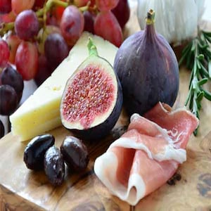 May include: A wooden cutting board with a variety of food items, including grapes, cheese, figs, prosciutto, and olives.